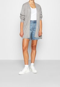Cardigan lavorato a maglia di un grigio chiaro con apertura frontale e maniche rilassate, abbinato a pantaloncini di denim. La modella indossa sneakers bianche e calzini alla caviglia.