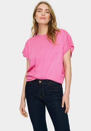 T-shirt rosa a maniche corte con scollatura rotonda, vestibilità leggermente ampia, abbinata a jeans blu scuro che evidenziano le cuciture e un dettaglio con bottone.