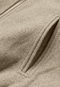 Beige ulljacka med en texturerad yta, dragkedjeslutning och två sidofickor med sydda kanter.