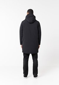 Parka negra acolchada con capucha, que presenta una longitud hasta media muslo, mangas largas y una textura suave. Lleva puesta sobre unos jeans negros y zapatillas.