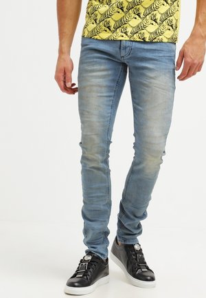Homme portant un jean skinny bleu clair associé à des baskets noires avec des détails de boucles argentées.