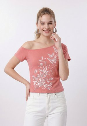 Χαμογελαστή γυναίκα με ροζ τοπ έξω από τους ώμους με άσπρο floral και πεταλούδες, φορώντας μεγάλα κρίκους σκουλαρίκια και άσπρο παντελόνι, ποζάροντας μπροστά σε άσπρο φόντο.