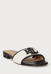 Muleit, joissa on kremi kanvaasi yläosa, mustat nahkakoristeet ja näkyvä musta logo. Litteä pohja, avoin varvas ja matala lohko korko.