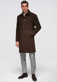 Ruskea villakangastakki lovikauluksella, napillinen suljenta ja sivutaskut, yhdistetty harmaisiin ruutuhousuihin ja ruskeisiin mokkanahkaisiin nilkkureihin.