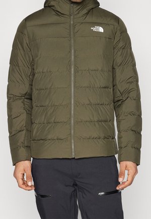 Homme portant une veste matelassée North Face vert olive avec capuche et un pantalon noir avec poches zippées.