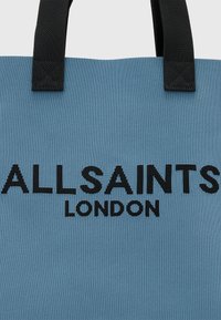 Jasnoniebieska torba na ramię z teksturowanej tkaniny, z czarnymi uchwytami; zawiera wyraźny czarny napis: "ALLSAINTS LONDON."