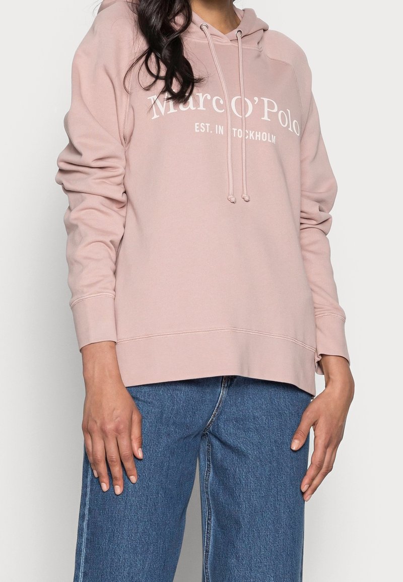 Felpa rosa realizzata in tessuto morbido, con cappuccio regolabile, logo frontale e polsini a coste, abbinata a jeans a gamba larga blu.