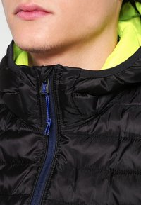 Nahaufnahme des unteren Gesichts und des Halses einer Person, die eine schwarze Daunenjacke mit einem neonfarbenen gelben Innenfutter der Kapuze und einem blauen Zipper-Zug trägt.