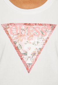 Camiseta de algodón blanca con un logo triangular de "GUESS" en rosa, con patrones florales, acentos de rhinestones y el texto "LAVADO".