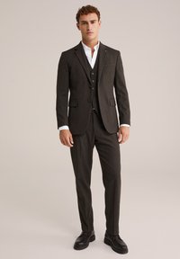 Tmavo šedý oblek na mieru so slim-fit nohavicami, trojbuttonovou vestou a bielou košeľou. Má jemnú látku a čierne spoločenské topánky.