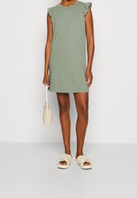 Robe verte à manches courtes avec détails à volants, réalisée en tissu léger. Accessoirisée d'un sac beige et de sandales plates froncées.