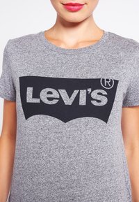 Kobieta ubrana w szary podkoszulek Levi's z czarnym nadrukiem logo, pokazana od ramion do górnej części klatki piersiowej na białym tle.