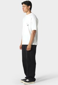 Valkoinen lyhythihainen t-paita, jossa on rintakuva, yhdistettynä löysiin mustiin housuihin ja mustiin tennareihin, joissa on valkoiset pohjat.