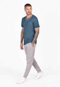 T-shirt in teal con effetto slub, dotato di taschino sul petto, abbinato a joggers grigi chiari; il modello indossa sneaker bianche, presentando un outfit casual.