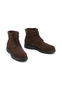Par de botas de ante marrón con cordones hasta el tobillo y cuellos acolchados, con suelas de goma negra, sobre un fondo blanco.