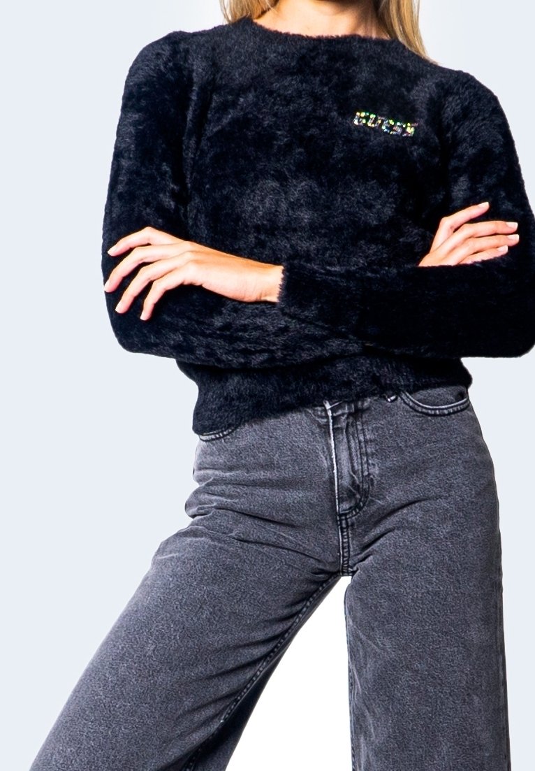 Personne portant un pull noir en matière duveteuse, bras croisés et jeans gris taille haute sur un fond clair uni.
