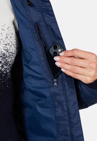 Mørkeblå pufferjakke med en lynlåslomme, hvor en hånd indsætter en mørk smartphone. Glat tekstur og letvægtsmateriale.