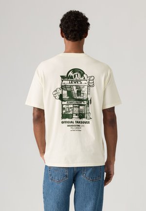 Homme portant un t-shirt crème avec un graphisme vert représentant la devanture d'un magasin, des yeux de dessin animé et un texte comprenant "Levi's", "Always Fresh" et "Official Takeover."