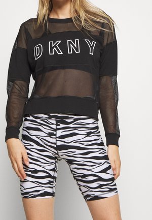 Osoba nosząca czarną siateczkową bluzkę z długim rękawem DKNY w połączeniu z wysokim stanem czarno-białych spodenek kolarskich w print zebry.