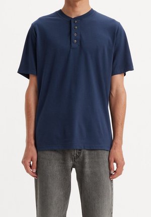 Man wearing navy blue short-sleeve henley shirt with four buttons, paired with faded gray jeans against a white background.