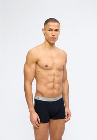 Γυμνασμένος άνδρας με κοντά μαλλιά που φοράει μαύρα μπόξερ Tommy Hilfiger, στέκεται μπροστά σε απλό φωτεινό φόντο.