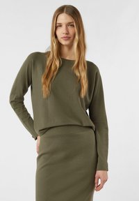 Sweter w kolorze oliwkowej zieleni, wykonany na drutach, z okrągłym dekoltem i długimi rękawami, zestawiony z dopasowaną spódnicą w tym samym kolorze. Gładka faktura i minimalistyczny design.