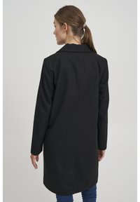 Cappotto lungo nero in misto lana, con colletto a punta e taglio dritto. Tessuto liscio, spacco sul retro e cuciture minime.
