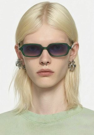 Personne aux cheveux blonds lisses, portant des lunettes de soleil rectangulaires vert foncé, de grosses boucles d'oreilles argentées et un haut vert clair.
