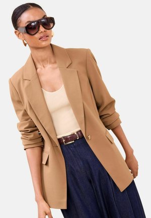 Brauner, maßgeschneiderter Blazer mit Schulternaht, getragen über einem beigen Oberteil und dunkelblauen, weit geschnittenen Hosen. Mit großen schwarzen Sonnenbrillen und einem Gürtel.