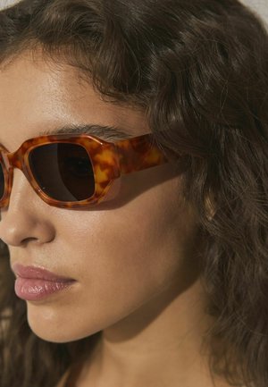 Göndör sötét hajú nő barna teknőspáncél mintás téglalap alakú napszemüvegben, közelről, profilból ábrázolva.