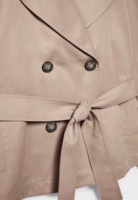 Manteau beige croisé avec de gros boutons foncés et une ceinture en tissu nouée autour de la taille.
