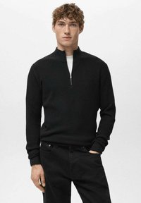 Czarny, prążkowany sweter z dzianiny z półgolfem na zamek. Posiada długie rękawy i dopasowane mankiety. Noszony z ciemnymi spodniami na jednolitym tle.