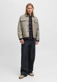Hellgraue Lederjacke mit zwei Vordertaschen über einem dunkelkarrierten Hemd. Dunkelblaue weite Jeans und bequeme beigen Slipper runden den Look ab.
