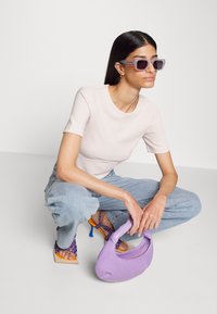 Top rosa claro com texturas, jeans azuis largos, mala roxa com uma silhueta curva e sapatos de salto alto coloridos com tiras.