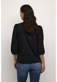 Svart topp med texturerade, spetsärmar i en chevronmönster. Enkel design, lättviktig tyg, ledig passform. Bakåtsidan visas.