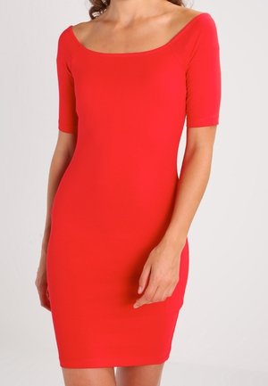 Femme portant une robe rouge ajustée à manches courtes et à encolure large et arrondie, debout devant un fond clair uni.