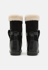 Bottines en suède noir avec un col en fourrure beige duveteuse, présentant une boucle sur le côté et des perforations décoratives sur le tige.