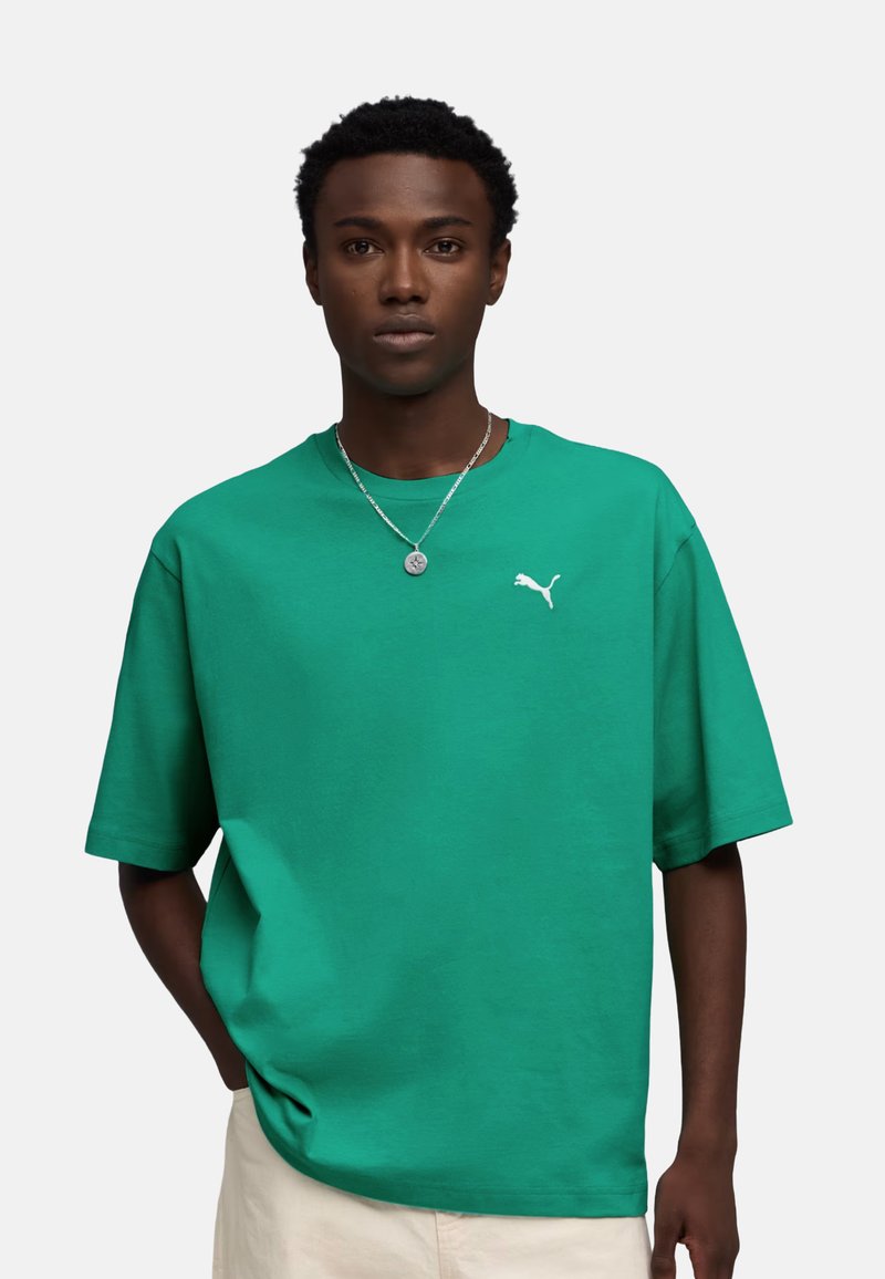 Jeune homme portant un t-shirt Puma vert vif et un pantalon beige, debout avec une main dans la poche devant un fond uni.