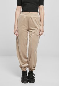 Beige velour joggerbyxor med elastisk midja; smalnade muddar. Bärs med svarta sneakers. Slät textur, avslappnad passform. Enkelt mönster.
