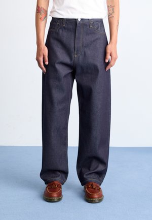 Dark blue denim pants with a loose fit, featuring yellow stitching and a button closure, paired with brown shoes.