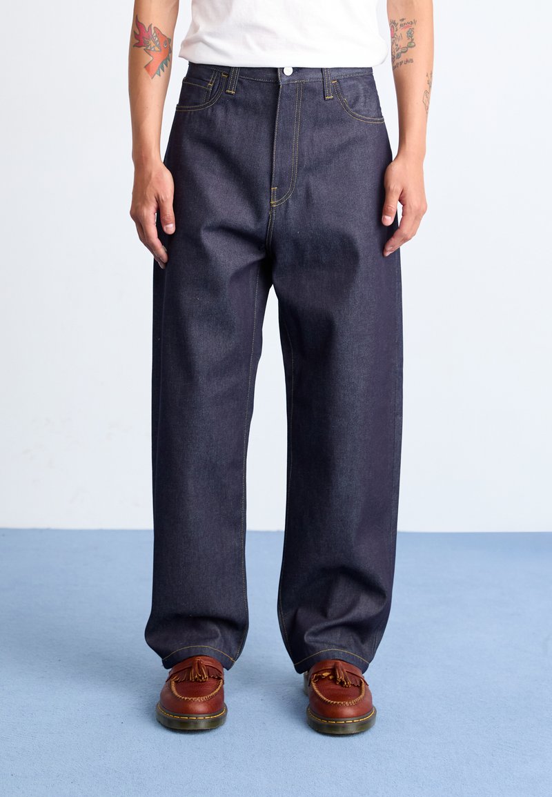 Dark blue denim pants with a loose fit, featuring yellow stitching and a button closure, paired with brown shoes.