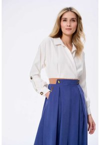 Biała bluzka z długim rękawem, z kołnierzykiem i zapinanymi mankietami, w połączeniu z niebieską spódnicą o wysokim stanem z fałdami z przodu i złotymi guzikami.