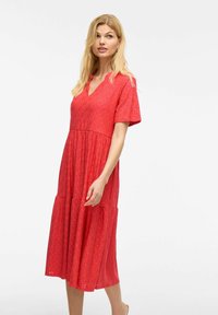 Robe mi-longue en coton rouge avec des manches courtes, décolleté en V, motif œillets, jupe superposée et coupe décontractée. Texture lisse et design léger.