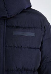 Námořnická modrá péřová bunda s kapucí a strukturovaným povrchem. Obsahuje horizontální prošívání a nášivku s logem Calvin Klein na prsou.