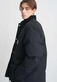 Chaqueta de plumas negra con cuello de pana, cierre de botones a presión y bolsillo frontal. Presenta una textura acolchada y un corte holgado.