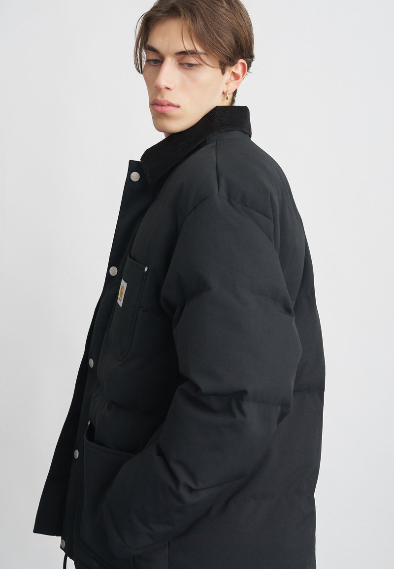 Chaqueta de plumas negra con cuello de pana, cierre de botones a presión y bolsillo frontal. Presenta una textura acolchada y un corte holgado.