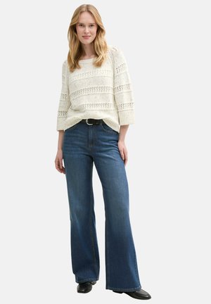 Pull en mailles crème avec des détails ajourés, manches trois-quarts, assorti à un jean en denim bleu évasé et des bottines noires.