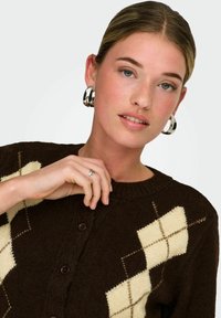 Cardigan en maille marron foncé avec un motif argyle crème, col rond et fermeture boutonnée à l'avant. Le modèle porte des boucles d'oreilles créoles argentées.