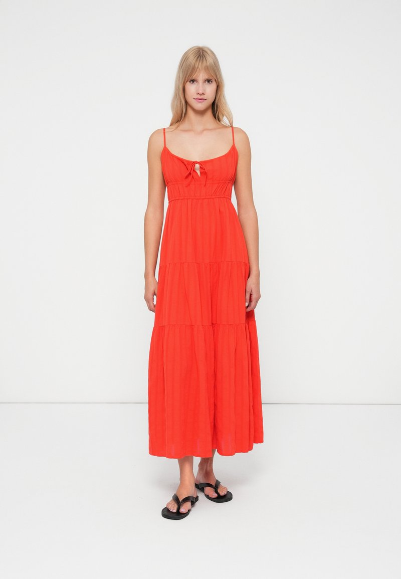 Vrouw staat wearing een mouwloze rode maxi-jurk met dunne banden en zwarte slippers tegen een eenvoudige witte achtergrond.