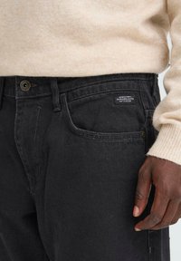 Pantaloni din denim negri cu o croială ușor tapered, având un buton metalic, nituri, buzunare frontale și o etichetă pe buzunarul din spate cu branding.